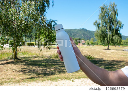Man holding in the hand white thermo stainless bottle with refreshing water on the nature. Man holding in the hand white thermo stainless bottle with refreshing water on the nature. 126608879