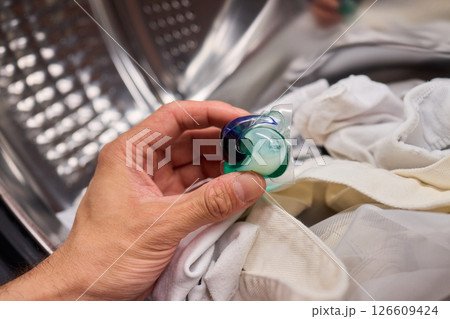 A hand is holding a colorful laundry pod as it is placed into a washing machine for cleaning 126609424