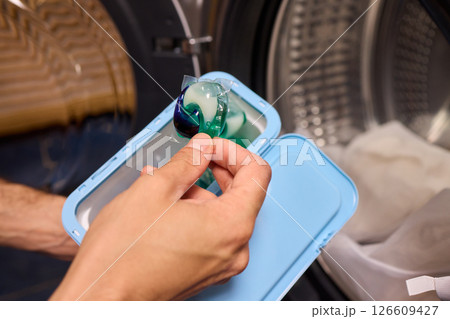 A hand is holding a colorful laundry pod as it is placed into a washing machine for cleaning 126609427