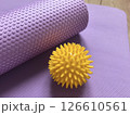 Sports equipment shown close-up: yellow massage ball and purple myofascial release roller behind it against the background of yoga, pilates and fitness mat lying on wooden floor. Fitness and health 126610561