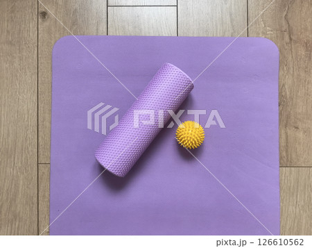 Top view of sports equipment: yellow massage ball, purple myofascial release roller and fitness yoga mat lying on wooden floor. Fitness and healthy lifestyle equipment 126610562