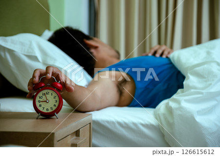Sleepy man lying in bed reaching out to turn off a ringing red alarm clock, representing morning tiredness and reluctance to wake up Sleepy man lying in bed reaching out to turn off a ringing red alarm clock, representing morning tiredness and reluctance to wake up 126615612