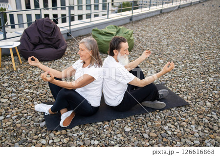 Senior couple performing relaxing meditation seated back to back in lotus pose outdoors. Man and woman wearing white and black activewear. Balanced lifestyle and harmony concept in urban environment. Senior couple performing relaxing meditation seated back to back in lotus pose outdoors. Man and woman wearing white and black activewear. Balanced lifestyle and harmony concept in urban environment. 126616868