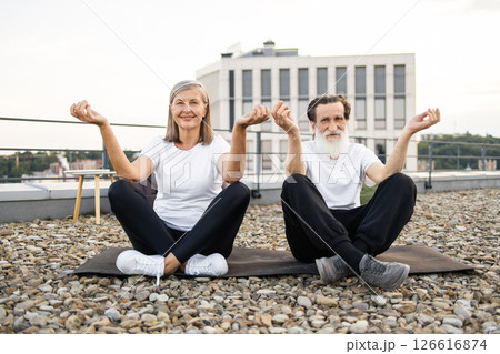 Senior man and woman performing yoga meditation in lotus pose on urban rooftop. Relaxation mindfulness wellness and bonding activities for older adults embracing active and healthy lifestyles outdoors 126616874