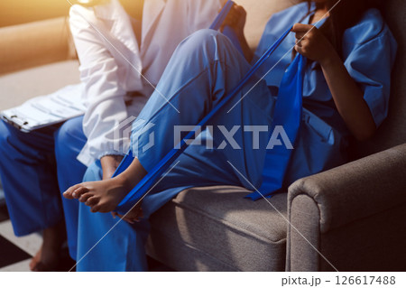 Confident physical therapist helps patient use resistance band stretching out his leg in clinic room. 126617488