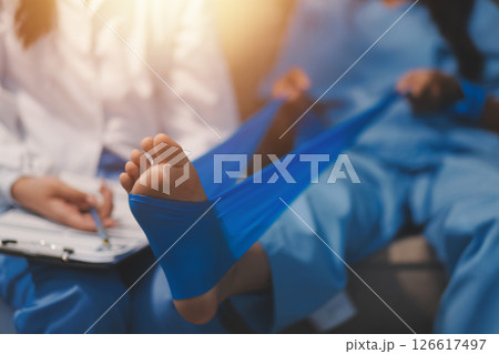 Confident physical therapist helps patient use resistance band stretching out his leg in clinic room. 126617497