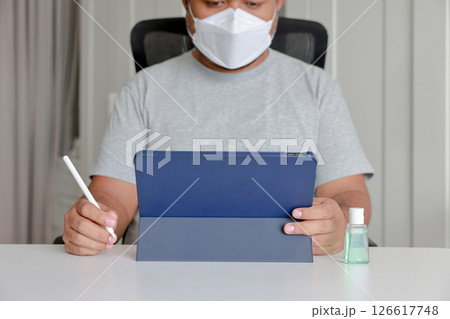 Man using tablet at home with hand sanitizer and face mask on table with stylus, concept of remote work, hygiene, and safety during pandemic or flu season. Man using tablet at home with hand sanitizer and face mask on table with stylus, concept of remote work, hygiene, and safety during pandemic or flu season. 126617748
