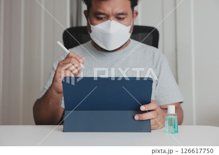 Man wearing face mask working on tablet with stylus at home, hand sanitizer on desk, concept of remote work, digital technology, and hygiene during pandemic or flu prevention. Man wearing face mask working on tablet with stylus at home, hand sanitizer on desk, concept of remote work, digital technology, and hygiene during pandemic or flu prevention. 126617750