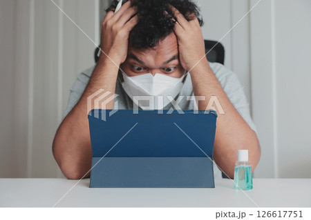 Worried man wearing mask looking shocked at tablet screen during pandemic, sitting at desk with hand sanitizer, stress and anxiety concept, quarantine life, remote work pressure. Worried man wearing mask looking shocked at tablet screen during pandemic, sitting at desk with hand sanitizer, stress and anxiety concept, quarantine life, remote work pressure. 126617751