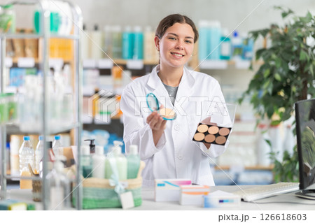 Professional young woman pharmacist standing at prescription counter and offering cosmetics for makeup and beauty products in household chemicals store or pharmacy 126618603