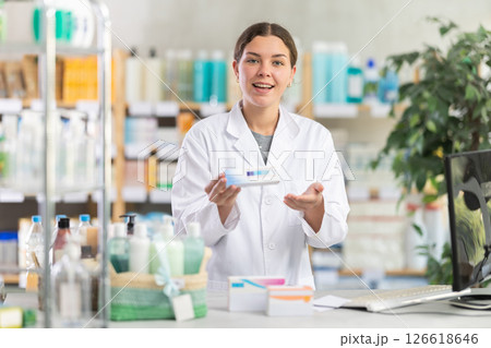 Skilled young woman pharmacist standing at prescription counter and offering medical ointment to clients at pharmacy shop Skilled young woman pharmacist standing at prescription counter and offering medical ointment to clients at pharmacy shop 126618646