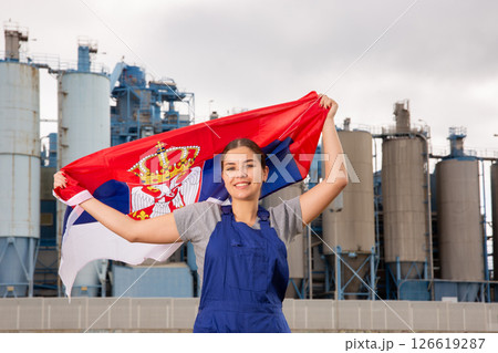 Happy young woman worker with flag of Serbia against background of factory 126619287