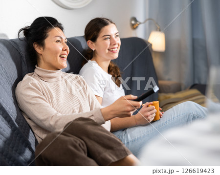 Two female friends watching TV while sitting on sofa at home 126619423
