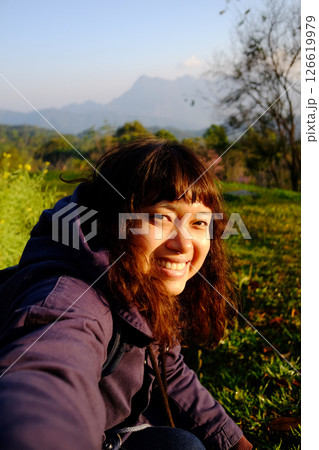 Traveler Asian woman wearing jacket and hood smiling in blooming wildflower on green lawn field with blue sky in winter on hill in Chiangmai of Thailand 126619979
