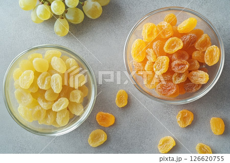 Top view of two glass bowls filled with golden raisins on gray stone background. Fresh grapes in frame. Natural texture, healthy snack presentation. Clean layout, bright light. Copy space 126620755