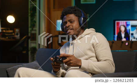 African american guy in modern apartment plays video games competition, relaxing and having fun. Person in multiplayer game on gaming console during leisure time at home. Camera A. African american guy in modern apartment plays video games competition, relaxing and having fun. Person in multiplayer game on gaming console during leisure time at home. Camera A. 126621203
