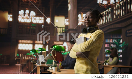 Portrait of cute teenager carrying textbooks and study materials in the library, preparing for a learning session to pass her exams. Young schoolgirl using books and academic resources. Camera A. 126621221