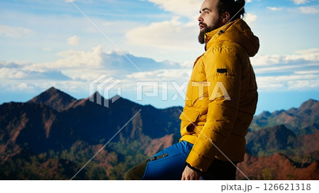 Adventure seeker gazes at the valley and mountain peaks ahead, feeling the freedom of the open wilderness. Young man fully equipped enjoying high altitude outdoor exploration. Camera A. Adventure seeker gazes at the valley and mountain peaks ahead, feeling the freedom of the open wilderness. Young man fully equipped enjoying high altitude outdoor exploration. Camera A. 126621318