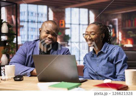 Black man and woman sits together in their cozy apartment space, discussing digital marketing strategy on a laptop. Supporting each other in their remote work and freelancing journey. 126621328