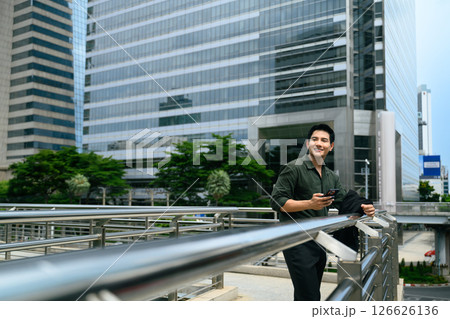 Smiling businessman leaning on railing in an urban area with smartphone in hand Smiling businessman leaning on railing in an urban area with smartphone in hand 126626136
