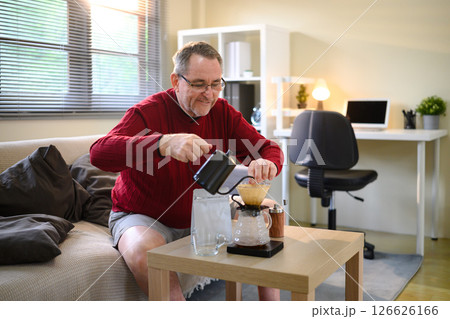 Mature male enjoying a slow coffee brewing process with a kettle and dripper in living room Mature male enjoying a slow coffee brewing process with a kettle and dripper in living room 126626166