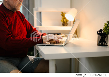 Close up of elderly man using laptop at minimalist desk under desk lamp Close up of elderly man using laptop at minimalist desk under desk lamp 126626184