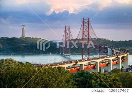 Scenic view of Lisbon iconic 25 de Abril Bridge over Tagus River with Cristo Rei or Christ the King statue in background at sunset. City traffic, lush greenery, tranquil river, famous landmarks, red Scenic view of Lisbon iconic 25 de Abril Bridge over Tagus River with Cristo Rei or Christ the King statue in background at sunset. City traffic, lush greenery, tranquil river, famous landmarks, red 126627467