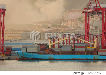 Blue container ship docked at Lisbon port, red cranes and stacks of shipping containers on deck, city buildings and church spire in evening haze and fire smoke in background. Risk of fires in summer 126627476