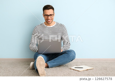 Caucasian man in striped shirt sitting on floor using laptop with notebook nearby against blue wall. concept of technology, casual work environment, modern lifestyle, productivity. 126627578