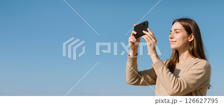 Young caucasian woman photographing outdoors with smartphone against clear blue sky. concept of outdoor photography, technology, travel, creative expression, Copy space, Wide banner 126627795