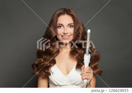 Good-looking young model woman with long curly wavy hairstyle showing hot curling tongs iron 126629055