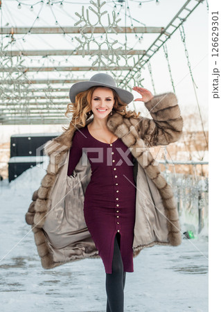 Pretty brunette woman walking on snow city street wearing hat red dress and fur coat Pretty brunette woman walking on snow city street wearing hat red dress and fur coat 126629301