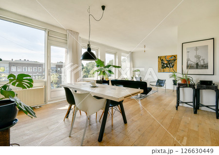 A stylish living room featuring a wooden table, minimalist furniture, and abundant natural light from large windows with a view of greenery outside. 126630449