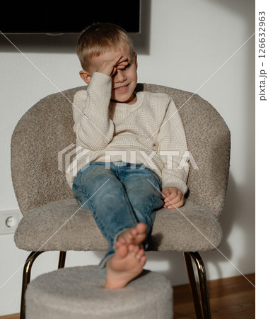 A 4-year-old boy sits in front of the window on a beige chair in the living room with sockless legs stretched forward and lying on a small chair with the sun shining directly in his face. High quality 126632963