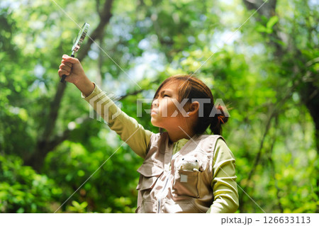 A happy little girl is exploring in the forest and looking for insects with a magnifying glass. Adventure tourism and learning concept. A happy little girl is exploring in the forest and looking for insects with a magnifying glass. Adventure tourism and learning concept. 126633113