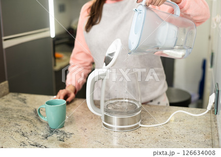 A woman in a bright kitchen pours water from a pitcher of filtered water into an electric kettle to prepare a hot drink. 126634008