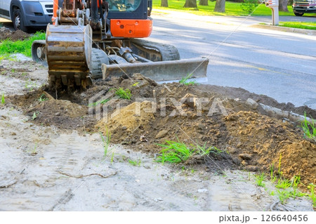 Excavator digging up soil along roadside for infrastructure development in residential neighborhood. Excavator digging up soil along roadside for infrastructure development in residential neighborhood. 126640456
