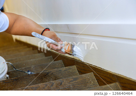 Hand holding brush applies white paint to trim near wooden stairs in home improvement effort. 126640464