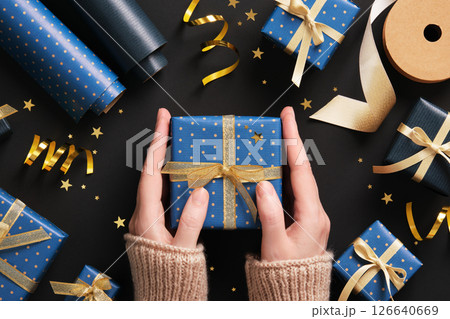 Hands hold out a blue gift box with gold ribbon surrounded by gift wrapping supplies on a dark background 126640669