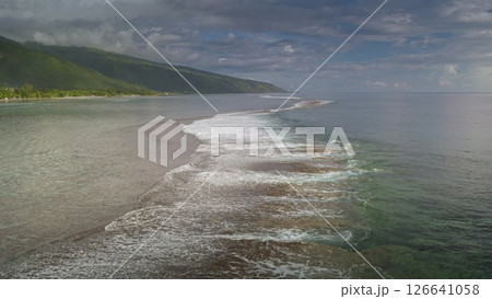 Stunning aerial view of waves gently breaking on a shallow reef, extending from a lush tropical island coastline under a dramatic cloudy sky Stunning aerial view of waves gently breaking on a shallow reef, extending from a lush tropical island coastline under a dramatic cloudy sky 126641058