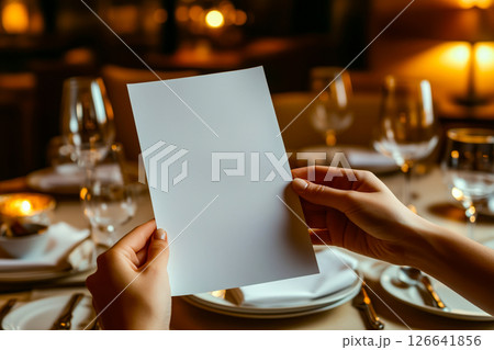 Hands holding a blank white menu card with an elegant dining table setup, glowing candlelight, and refined glassware in a cozy restaurant ambiance 126641856
