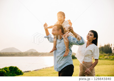 Asian family child girl piggyback riding back on father raising hands like flying outdoor nature park with mother, Happy parent daughter having fun together, Tourist lifestyle family leisure activity 126643840