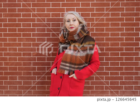 Gray-haired woman of sixty years old in a red coat and a terracotta scarf near a brick wall Gray-haired woman of sixty years old in a red coat and a terracotta scarf near a brick wall 126645645