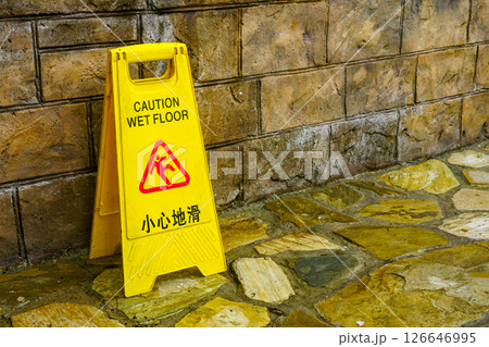 Yellow caution wet floor sign with English and Chinese text placed near a stone wall on wet surface Yellow caution wet floor sign with English and Chinese text placed near a stone wall on wet surface 126646995