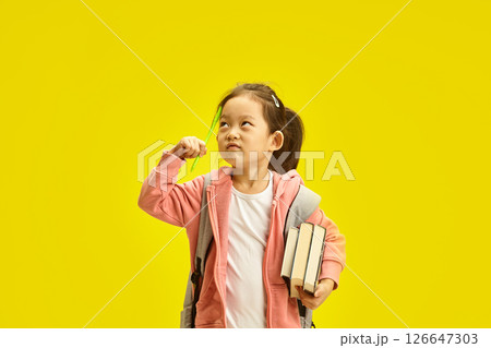 Curious Korean Preschool kid girl with Books and Pencils stands concentrating on scratching herself temple with a pen, looking up thoughtfully while standing on a yellow background 126647303