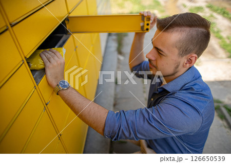 A Young Man Happily Collecting a Package from a Bright Yellow Mailbox in His Neighborhood 126650539