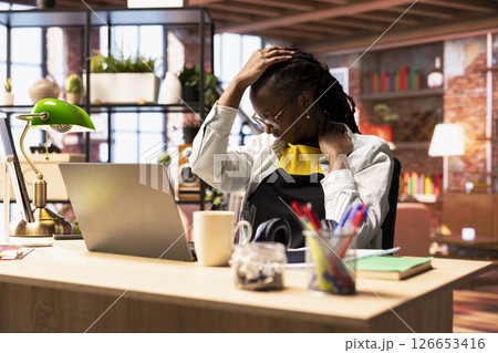 Fatigued woman in home office stretching sore neck after sitting at desk too long, taking break. Exhausted freelancer in apartment doing muscle exercises to relieve painful stinging muscle spasm 126653416
