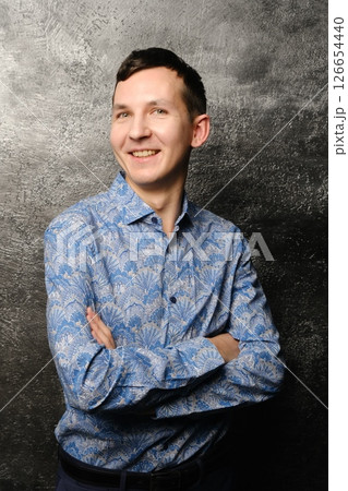 A young man with a bright smile in a stylish blue patterned shirt stands against a dark background A young man with a bright smile in a stylish blue patterned shirt stands against a dark background 126654440