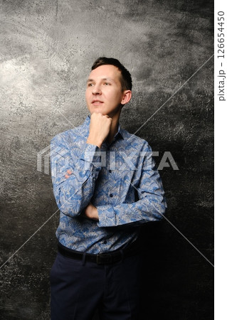 A Thoughtful Young Man in a Stylish Blue Shirt Posing Elegantly Against a Dark Background A Thoughtful Young Man in a Stylish Blue Shirt Posing Elegantly Against a Dark Background 126654450
