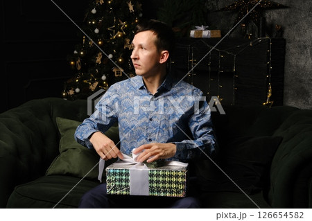 A Joyful Man Unwrapping a Gift Next to a Beautifully Decorated Christmas Tree with Bright Lights 126654582
